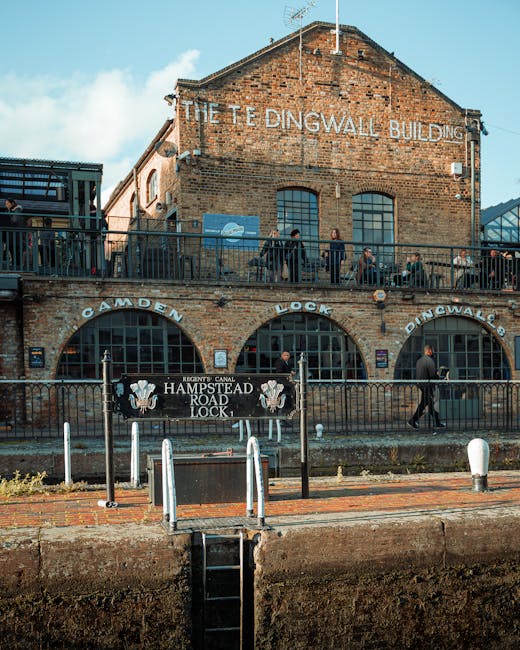 A multi-storey brick building labeled 'The Tedingwall Building' with large arched windows on the ground floor, above which are smaller rectangular windows. A balcony runs along the second floor, with several people standing and sitting, some leaning on the black metal railing. In front of the building, there are black metal fences and a sign that reads 'Hampstead Road Lock,' indicating a canal lock nearby, with a lock opening visible at the bottom of the image leading to the water's edge. The scene is set outdoors during daytime with a partly cloudy sky, and the area appears to be part of a historic or scenic site in Hampstead. This setting may be relevant for house removals or transport services in the Hampstead area, as managed by Man and Van Hampstead, highlighting elements of moving logistics and planning.