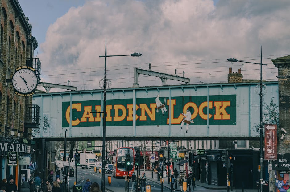 A busy London street scene showing a large freight train bridge with a green and yellow Camden Lock sign painted on the side. The bridge spans over a pedestrian area with people walking and waiting at bus stops. Trees and streetlights are visible, along with a red double-decker bus and various shopfronts on the street level. To the left, a building with a clock and a small balcony can be seen, and to the right, a red sign and additional street furniture are present. The scene captures an active urban environment typical of a central London area, relevant to house removals and furniture transport services such as those provided by Man and Van Hampstead, which operate in this locale for home relocations and moving logistics.