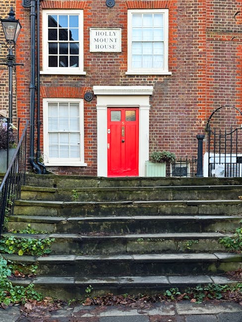 A multi-storey brick building labeled 'The Tedingwall Building' with large arched windows on the ground floor, above which are smaller rectangular windows. A balcony runs along the second floor, with several people standing and sitting, some leaning on the black metal railing. In front of the building, there are black metal fences and a sign that reads 'Hampstead Road Lock,' indicating a canal lock nearby, with a lock opening visible at the bottom of the image leading to the water's edge. The scene is set outdoors during daytime with a partly cloudy sky, and the area appears to be part of a historic or scenic site in Hampstead. This setting may be relevant for house removals or transport services in the Hampstead area, as managed by Man and Van Hampstead, highlighting elements of moving logistics and planning.