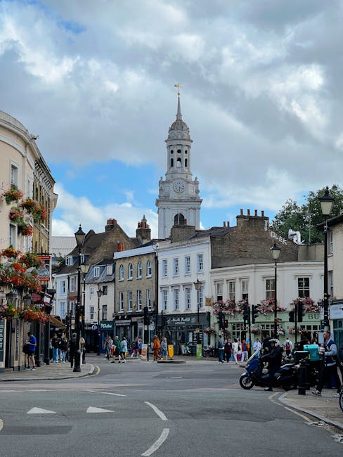 A street scene in Hampstead showing a busy intersection with pedestrians walking along the pavements and crossing the road. Several retail shops line the street, with some displaying signage and window displays. In the foreground, a motorcycle with two riders is waiting at the traffic lights, while others wait or walk nearby. The background features historic buildings with brick facades and a tall, ornate clock tower with a spire, set against a partly cloudy sky. The scene captures typical urban activity during daytime, with some people carrying shopping bags or engaged in conversation. This setting is consistent with an area where professional removals services, such as those offered by Man and Van Hampstead, could be involved in home relocation or furniture transport, highlighting the importance of efficient moving logistics in such busy urban environments.