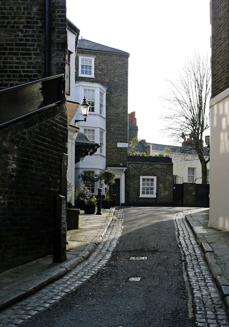 A narrow residential street in Hampstead with a cobbled pavement and curb sides, flanked by brick buildings on both sides. On the left, there is a black brick building with a small, black metal railing along the pavement, and a lamppost mounted on the wall near a white door. The right side features a white modern building with smooth exterior walls and a window, partially visible behind a white panel or barrier. In the background, there are trees with bare branches and additional buildings, indicating a typical urban area suitable for home relocation activities. The street appears quiet and well-maintained, with natural daylight illuminating the area, which supports the context of local house removals and furniture transport near Hampstead Heath.