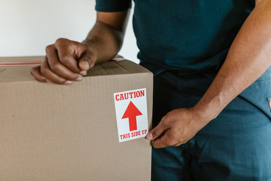A person wearing a dark shirt and blue trousers is holding a large cardboard moving box with both hands, with their left hand gripping the side and their right hand grasping the top. The box features a white label with red text and an arrow, reading 'CAUTION THIS SIDE UP,' indicating careful handling during the packing and moving process. The background shows an indoor setting, possibly inside a property, with neutral-colored walls. The scene emphasizes the careful handling of packed items in a home relocation, as might be coordinated by Man and Van Hampstead during furniture transport for house removals or commercial office moves. The image captures the loading process, illustrating the importance of proper packing, orientation, and transport of household or office belongings.