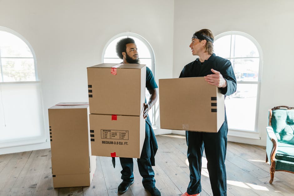 A man is indoors, lifting and carrying a medium-sized cardboard box labeled 'OFFICE' during a house or office move. He is wearing a beige t-shirt and grey trousers, with a focused expression. The room has white walls and a large window with sheer white curtains allowing natural light to fill the space. In the background, there is a metal shelving unit with a potted plant on top, and several other boxes, some labeled 'MEDIUM', are stacked on the wooden floor. The boxes are made of brown cardboard, some wrapped in plastic or packing tape, indicating packing and moving preparations. This loading process illustrates furniture transport and packing activities typical of professional removals, such as those offered by Man and Van Hampstead, involved in home relocation and small business moves.